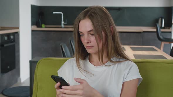 Shocked and Worried Young Woman Reading Bad News in Message on Her Smartphone While Sitting on Sofa