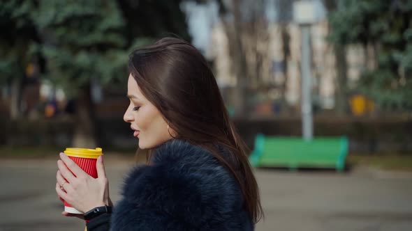 Pretty girl with hot drink outdoors. Attractive young woman in luxury fur coat enjoys coffee in the 