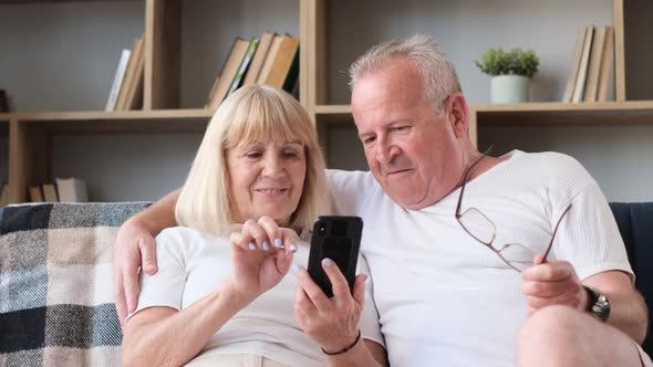 An Elderly Husband and Wife are Sitting on the Sofa in the Living Room and Mastering a Smartphone