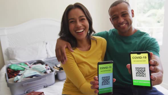 Happy biracial couple sitting on bed and showing smartphones with covid 19 passports on screens