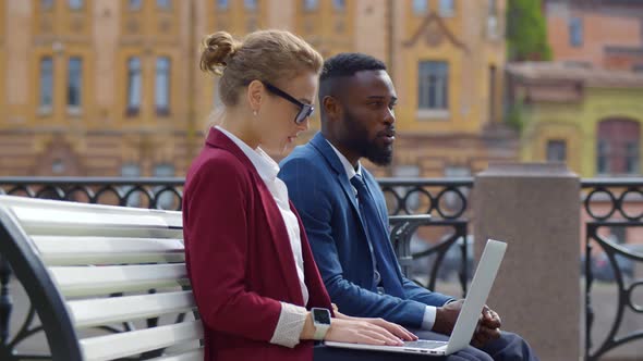 Side View of Multicultural Business Couple Working on Laptop Sitting on Bench Near Office.