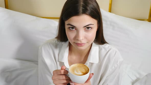 Portrait of Young Attractive Confident Woman Sitting on White Bedding Holding Cup with Coffee and