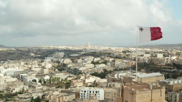 Slow Establishing Shot Passing By Flag of Malta Waving in Revealing City on Gozo Island with Cloudy