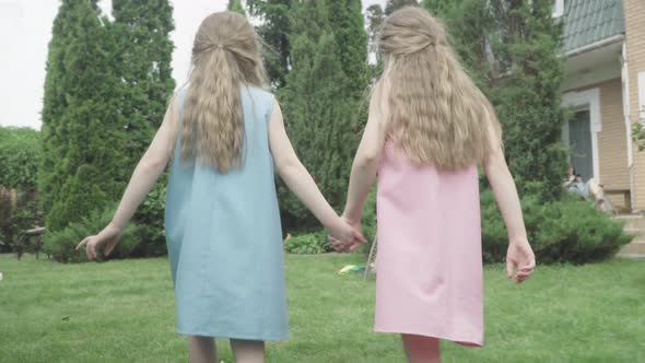 Camera Follows Happy Twin Sisters Running To Green Tree on Backyard. Back View of Joyful Caucasian