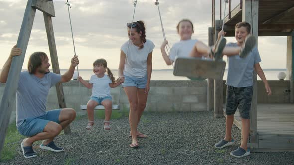 Happy Family Rolls Young Children on a Swing Outdoors at Sunset.