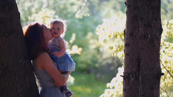 baby and mother are playing standing in the forest by a tree trunk in backlight