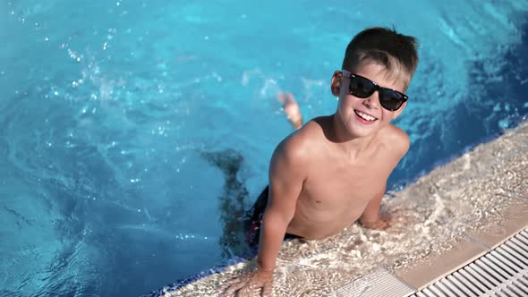 Cheerful Child Boy Tanned Skin Wearing Sunglasses Playing Legs Splashing Water at Swimming Pool