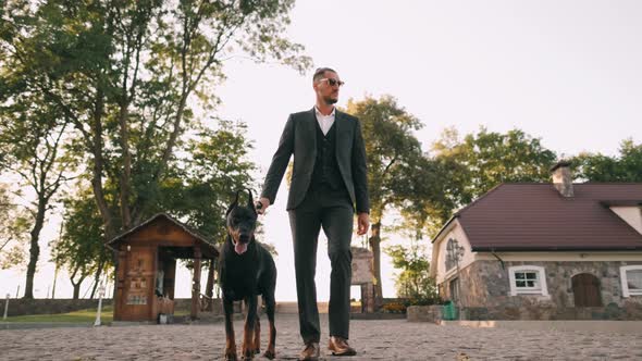 A Rich Businessman in a Business Suit Walks with a Large Doberman in the Park. Successful Bearded