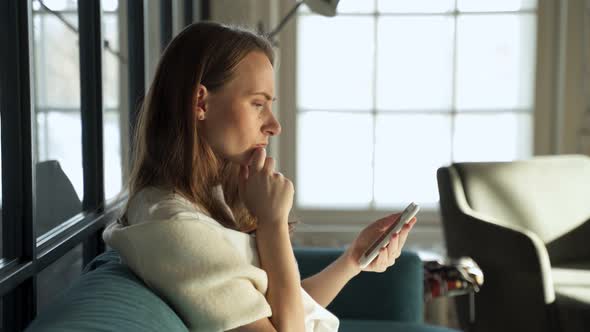 Serious Excited Young Woman Exchanges Text Messages Using a Mobile Phone While Sitting at Home on