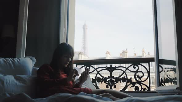 Beautiful Happy Blogger Woman Lying on Bed at French Balcony Window with Eiffel Tower View Using