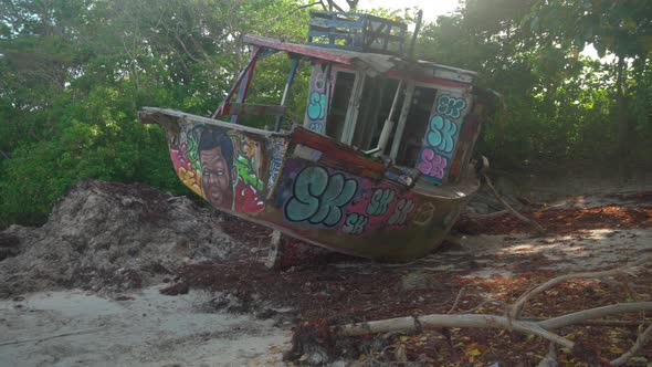 Graffitied Abandoned Fishing Shipwreck sits on Dirt Surrounded by Trees with Sunlight Gleaming throu