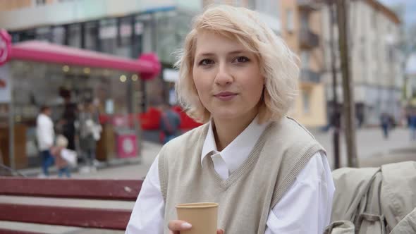 Young Blonde Business Woman in a Beige Vest and White Blouse Sits on a Bench in the Middle of the