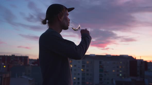 Young Man Drinks Wine and Look at Sunset Over City