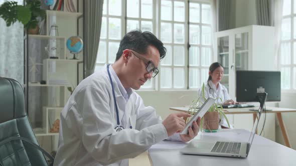 Asian Man Doctor Is Using Tablet While Work With Laptop Computer In Workplace. Medical Concept