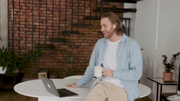 A Man Drinks Coffee at the Workplace and Talks on Wireless Headphones