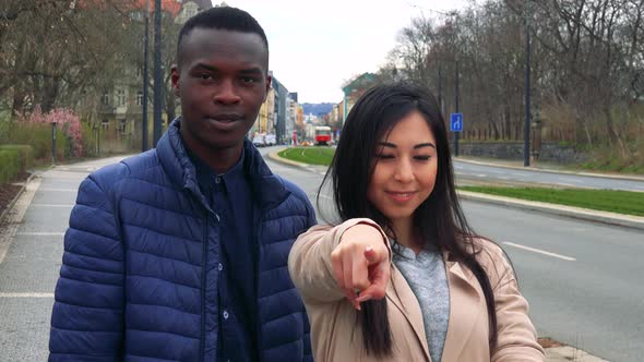 A Young Black Man and a Young Asian Woman Smile and Point at the Camera on a Sidewalk, an Urban Area