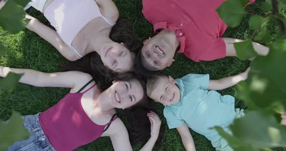 Happy Family Laying Down On A Green Grass And Looking Up Smiling In A Shade Of A Tree In The Summer