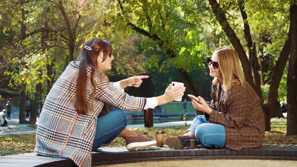 Stylish Girls Sitting Face to Face and Using Smartphones in Autumn Park