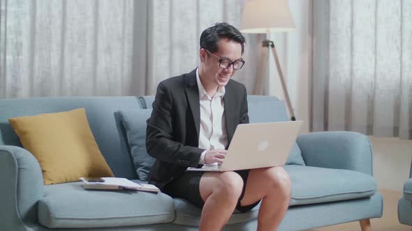Smiling Asian Businessman In Jacket And Shorts Sitting On Sofa And Typing On A Laptop At Home