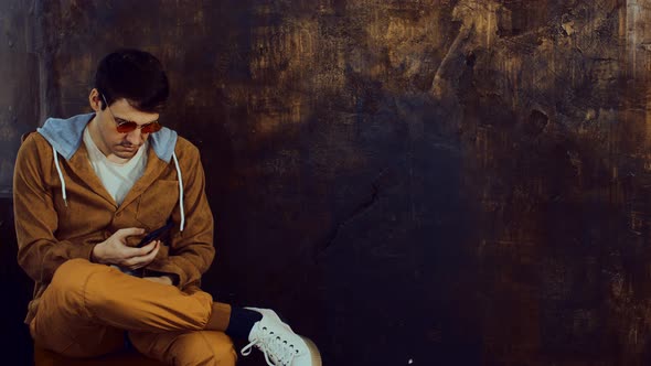 Stylish Man in Bright Glasses Sits on Chair Browsing Mobile Phone Near Shabby Brown Wall