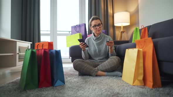 Happy Woman with Glasses Is Sitting on the Floor and Makes an Online Purchase Using a Credit Card