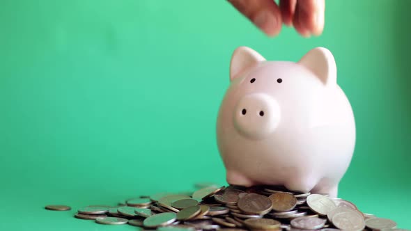 Man Putting Coins in Piggy Bank Standing in Pile of Coins on Green Background.