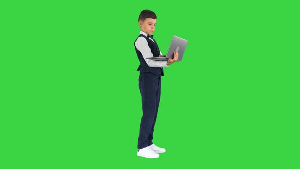 Concentrated Boy in a Bow Tie and Waistcoat Using Laptop Computer While Standing on a Green Screen