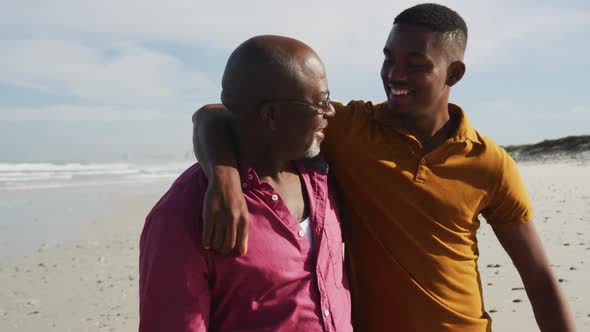 African american senior father and teenage son standing on beach embracing and talking