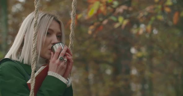 Woman Swinging While Drinking Hot Tea in the Middle of a Cold Park in Autumn. Young Woman Reminds