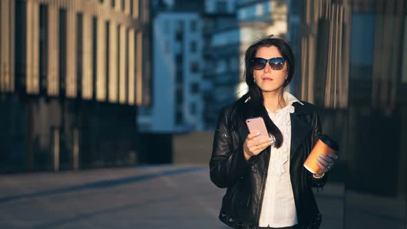 Young Attractive Businesswoman Walking with Smart Phone in the City with Office Buildings