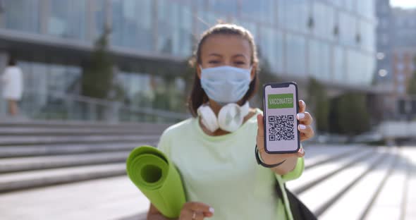 Sporty Woman in Safety Mask Looking at Camera and Showing Smartphone with Vaccination Certificate