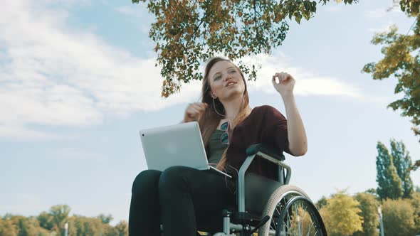 Happy Caucasian Woman in the Wheelchair Listening the Music on Laptop Under the Tree. Low Angle