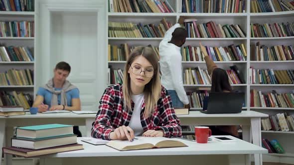 Girl in Glasses Sitting at the Table in Reading Room and Doing University Task