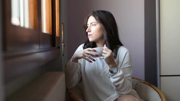 Young eastern woman at home sitting near window relaxing in her living room drinking coffee or tea