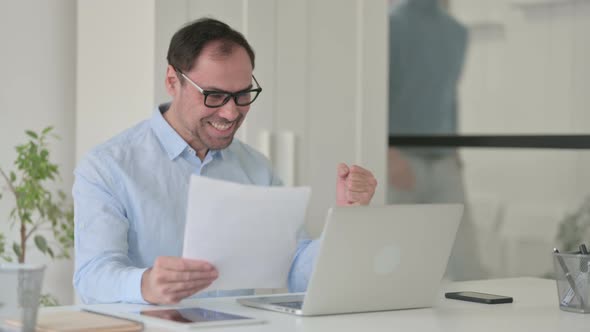 Successful Middle Aged Man Reading Documents While Using Laptop