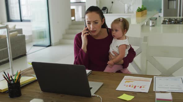 Caucasian mother holding her baby and talking on smartphone while working from home