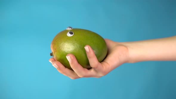 Mango Apple with Eyes in a Woman Hand Close Up. Apple Looks Around on a Blue Background. Vertical