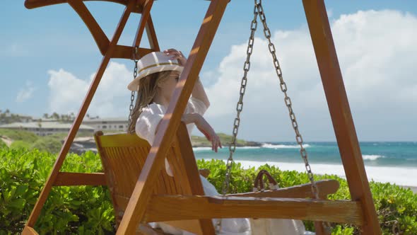 Happy Young Woman in Fashionable White Fedora Hat on Wooden Swing at Ocean Park