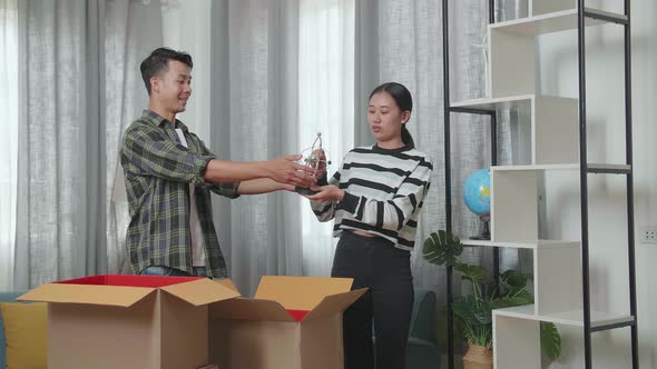Young Asian Couple Moving Into A New House Taking Stuff Out Of The Cardboard Box To Bookcase