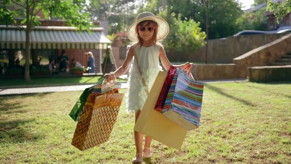 Little Fashion-girl with Shopping Bags After Visiting Expensive Boutiques Walks on Lawn and Smiles