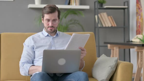 Young Man with Laptop Reading Documents on Sofa