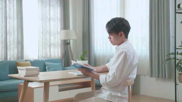 Side View Of Asian Man Student Reading A Book While Studying At Home