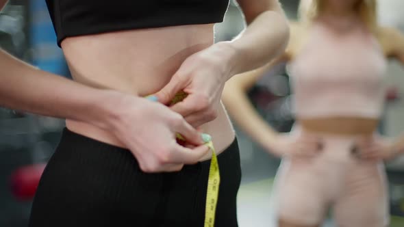 Unrecognizable Sportswoman Measuring Waist in Gym with Blurred Personal Fitness Trainer Standing at