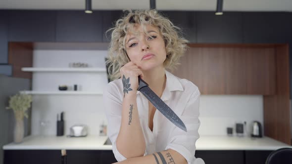 Portrait of Tattooed Blond Caucasian Woman Holding Kitchen Knife Looking at Camera with Serious