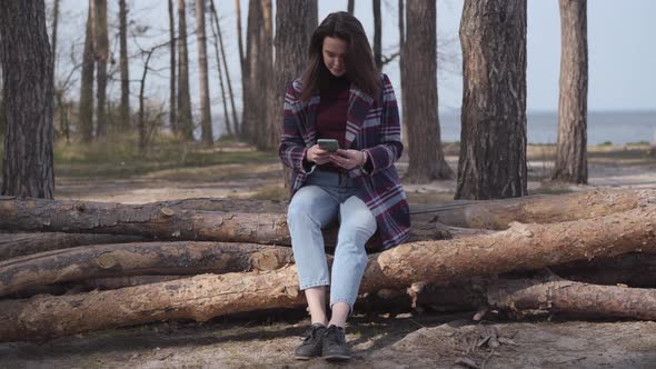 Happy Smiling Woman Sitting on Log in Forest and Using Smartphone. Brunette Caucasian Girl Resting