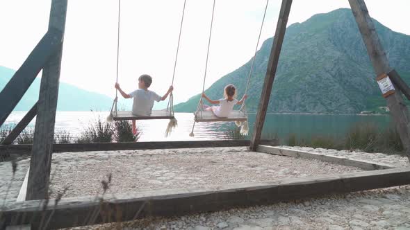 A Girl and a Boy Swing on a Swing Near the Sea with a Beautiful View of the Bay and Mountains