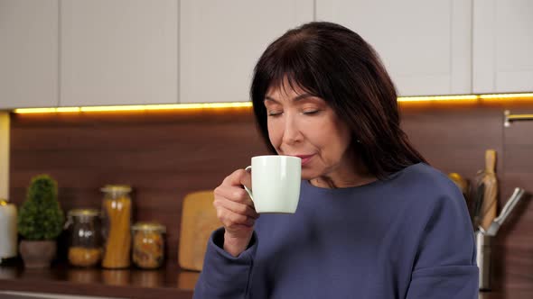 Aged Woman Drinking Delicious Warm Tea Coffee From Cup in Home Kitchen