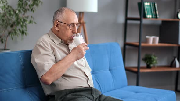 Happy Elderly Man Drinking Fresh Milk Glass Sitting on Comfortable Couch Enjoying Retirement at Home