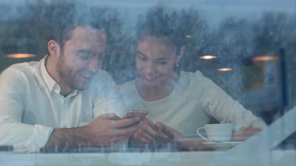 Happy Young Couple Sitting at Table Using Phone in Cafeteria