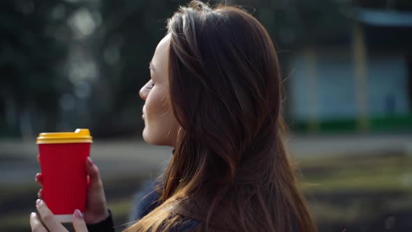 Beautiful brunette in the park. Long-haired girl in fur coat holds plastic cup outdoors. Camera move